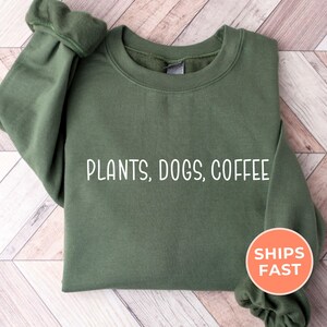 May include: Military green sweatshirt with the words "PLANTS, DOGS, COFFEE" in white. The sweatshirt has a crew neck and long sleeves. An orange circle with the words "SHIPS FAST" is on the right side.