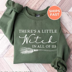 May include: A military green crewneck sweatshirt featuring white text that reads There's a little Witch in all of us, accompanied by a graphic illustration of a broomstick positioned horizontally beneath the printed lettering.