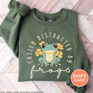 May include: Military green sweatshirt with the text "EASILY DISTRACTED BY frogs" and a cartoon frog illustration. The frog is light blue with large eyes, surrounded by yellow flowers. The sweatshirt has a crew neck and long sleeves.