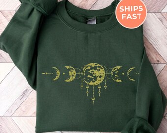 May include: Forest green sweatshirt featuring a gold moon phases design. The graphic includes a full moon and crescent moons connected by lines and dangling accents. The sweatshirt has a crew neck and long sleeves. The text "SHIPS FAST" is in a circle in the upper right corner.