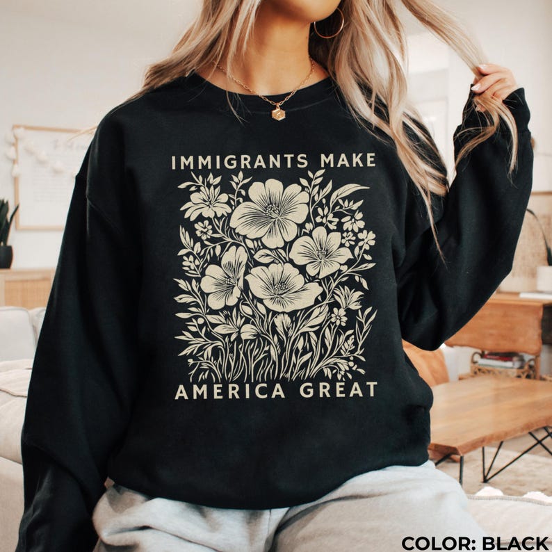 May include: Black sweatshirt with the text "IMMIGRANTS MAKE AMERICA GREAT" in white. The design features a floral illustration in cream, including large flowers and foliage. The sweatshirt is a solid black color.