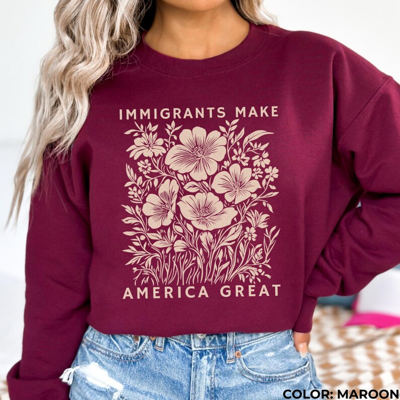May include: Maroon sweatshirt with a floral design and the text "IMMIGRANTS MAKE AMERICA GREAT" in a cream color. The design features large flowers and foliage. The sweatshirt is made of a soft material.