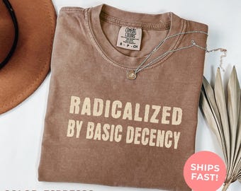 May include: Espresso-colored t-shirt with the text "RADICALIZED BY BASIC DECENCY" in a bold, off-white font. A silver necklace with a small pendant rests on the shirt. The shirt has a Comfort Colors tag.