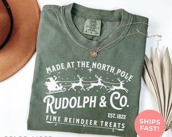 Könnte beinhalten: Ein moosgrünes Kurzarm-T-Shirt mit einer weißen Grafik von Santa im Schlitten, gezogen von Rentieren. Der Text lautet Made at the North Pole, Rudolph & Co. Est. 1822 und Fine Reindeer Treats. Das Shirt hat ein Comfort Colors Markenetikett.