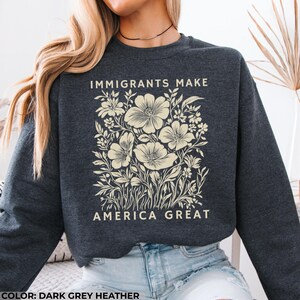May include: Dark grey heather sweatshirt with the text "IMMIGRANTS MAKE AMERICA GREAT" in white. The sweatshirt features a floral design in cream, with large flowers and detailed leaves. The overall style is casual and comfortable.