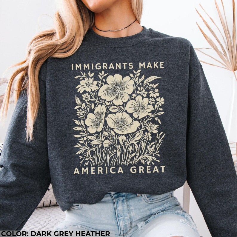 May include: Dark grey heather sweatshirt with the text "IMMIGRANTS MAKE AMERICA GREAT" in white. The sweatshirt features a floral design in cream, with large flowers and detailed leaves. The overall style is casual and comfortable.