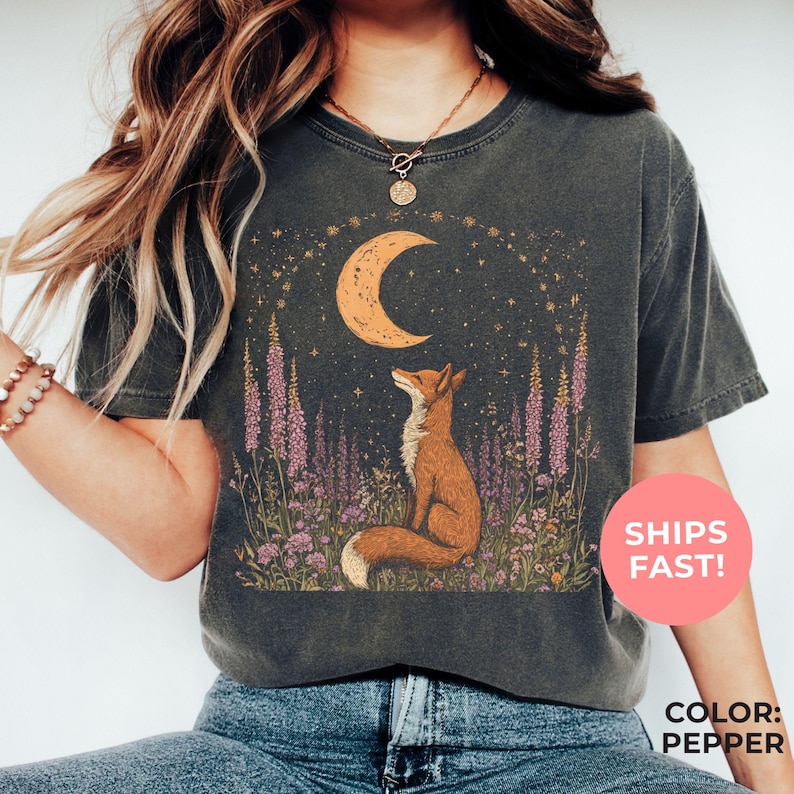 An pepper-colored shirt, illustrating a celestial fox nestled among flowers and gazing at the moon — good for nature lovers, stargazers, and those drawn to mystical woodland vibes.