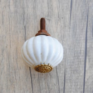 May include: White ceramic cabinet knob with a fluted design and a gold metal accent. The knob has a rusty metal screw.