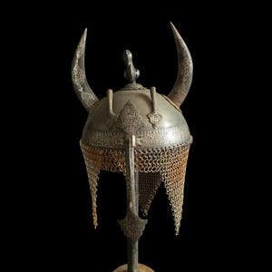Puede incluir: Un casco plateado antiguo con dos cuernos grandes y curvos. El casco presenta intrincados patrones grabados y una protección para el cuello de cota de malla. Una pequeña figura con forma de pájaro se encuentra en la parte superior del casco. El casco se exhibe sobre un soporte sobre un fondo negro.