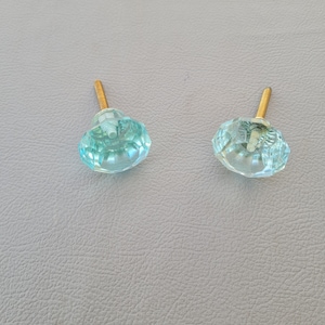 Könnte beinhalten: Zwei hellblaue Glas-Schrankknöpfe mit goldenen Metallschrauben. Die Knöpfe haben ein facettierten Design.