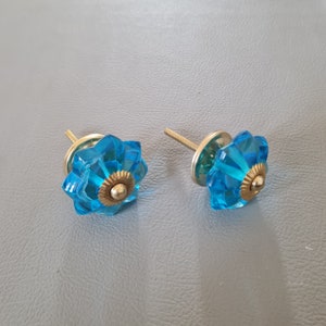 Könnte beinhalten: Zwei blaue Glas-Schrankknöpfe mit goldener Metallbasis. Die Knöpfe haben ein blumenartiges Design.