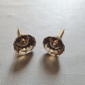Op de afbeelding: Twee bruine glazen kastknoppen met gouden metalen schroeven. De knoppen zijn gefacetteerd en hebben een geometrische vorm.