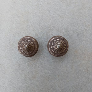 以下が含まれることがあります： 装飾的な花のデザインが施された銀色の金属製のボタンが2つ。ボタンは中央が盛り上がっており、わずかに摩耗した仕上げになっています。