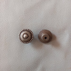 May include: Two silver-toned metal knobs. One knob has a decorative design with a raised center circle. The other knob is plain.