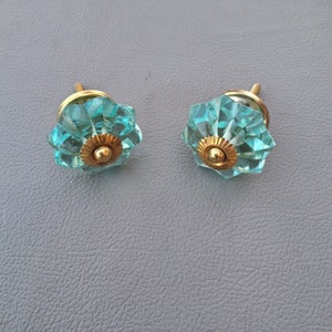 Puede incluir: Dos pomos de armario de cristal azul turquesa con detalles en metal dorado. Los pomos tienen un diseño facetado.