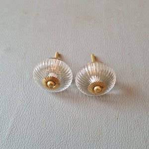 Könnte beinhalten: Zwei klare Glas-Schrankknöpfe mit goldener Metallakzente. Die Knöpfe haben ein geripptes Design.