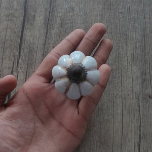 Può includere: Pomello per armadio a forma di fiore in ceramica bianca con centro marrone scuro e dettagli dorati. Il pomello è tenuto in mano su uno sfondo di legno. Questo hardware decorativo può essere utilizzato per cassetti o armadi.