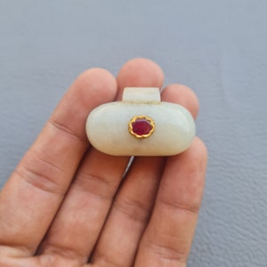 May include: A white oval pendant with a gold setting and a red gemstone. The pendant is held in a hand.