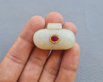 May include: A white oval pendant with a gold setting and a red gemstone. The pendant is held in a hand.