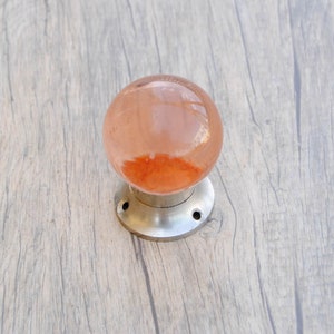 May include: A round, peach-colored glass door knob with a silver metal base and three screws.
