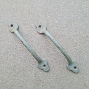 May include: Two vintage, light blue metal cabinet handles. Each handle has a curved bar and decorative ends with a single hole for mounting. The paint shows signs of wear, giving them a rustic appearance. The handles are set against a neutral gray background.