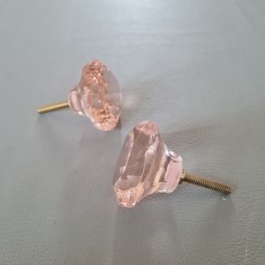 Könnte beinhalten: Zwei rosafarbene Glas-Schrankknöpfe mit goldenen Metallschrauben. Die Knöpfe sind diamantförmig.