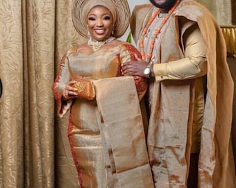 May include: A couple in traditional Nigerian attire. The woman wears a gold-colored dress with a matching head wrap and clutch. The man wears a gold-colored outfit with a matching hat and sunglasses. Both are adorned with coral beads.