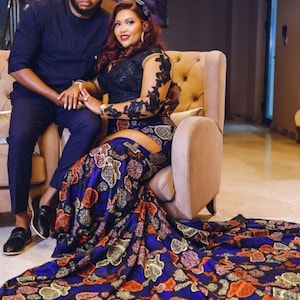 May include: A couple poses for a photo in a formal setting. The woman is wearing a black top with a long-sleeved lace overlay and a blue and orange patterned skirt. The man is wearing a dark blue shirt and black pants.