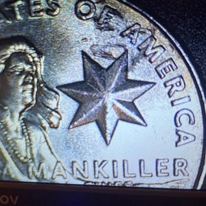 May include: Close-up of a silver coin with the words "STATES OF AMERICA" and "MANKILLER" visible. A seven-pointed star is in the center. The coin features a portrait of a Native American woman.