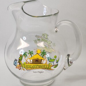 May include: A clear glass pitcher featuring a colorful graphic of a tropical hut, palm trees, a parrot, and a margarita glass. The text reads "It's 5 O'Clock Somewhere", "MARGARITAVILLE", and "Las Vegas" printed in green, yellow, and black ink.