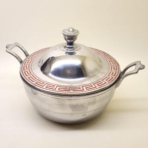 May include: A silver metal serving dish with a lid and handles. The lid features a red and white Greek key design around the edge. The dish has a round shape and a small knob on top of the lid.
