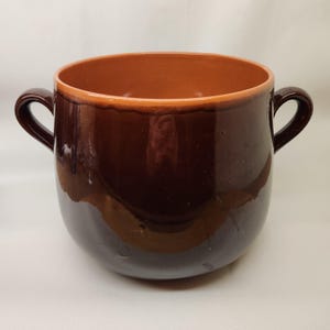 May include: A brown earthenware pot with two handles. The pot has a rounded bottom and a wide opening. The pot is glazed in a dark brown color with a lighter brown rim.
