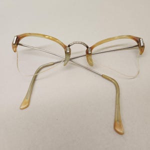 May include: Vintage eyeglasses with a half-rim design. The frames feature a light brown and silver color scheme, with decorative accents on the bridge and temples. The lenses are clear, and the arms have a curved shape for comfortable wear.