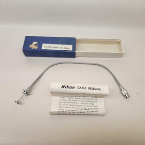 May include: A Nikon cable release and wire release set. The set includes a silver-colored flexible cable, a blue box labeled "Nikon Wire Release," and a white paper with "Nikon Cable Release" printed on it. The paper contains instructions.