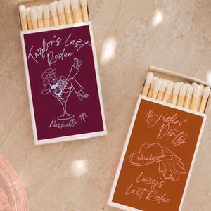 May include: Two rectangular matchboxes with white wooden matches. The dark maroon box features a white line drawing of a person in a martini glass with the text Taylor's Last Rodeo Nashville, TN. The orange box shows a cowboy hat and text.