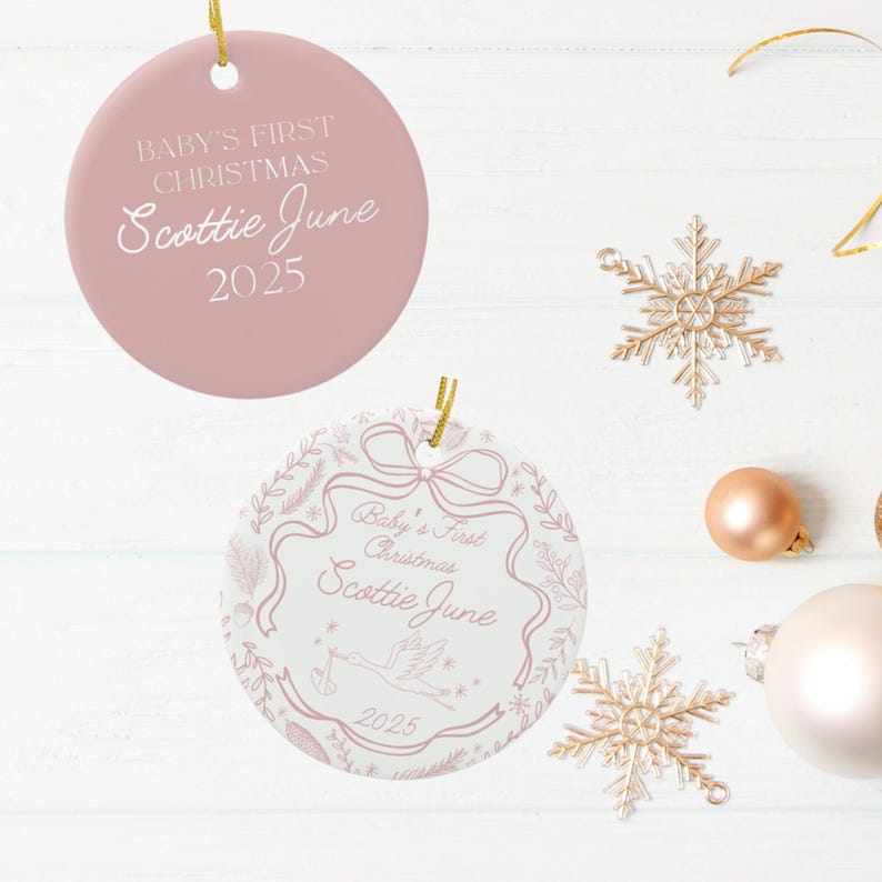 Two round ceramic ornaments, one solid pink with white text and one white with pink text and illustrations, are displayed with gold snowflake ornaments and a rose gold ball ornament on a white surface.