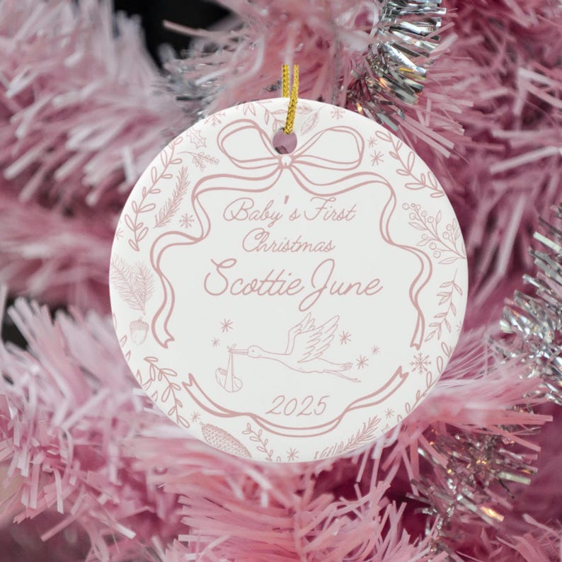 A round, white ceramic ornament with pink text and illustrations, reading "Baby's First Christmas Scottie June 2025" and showing a stork carrying a baby, hangs on a pink tinsel Christmas tree.