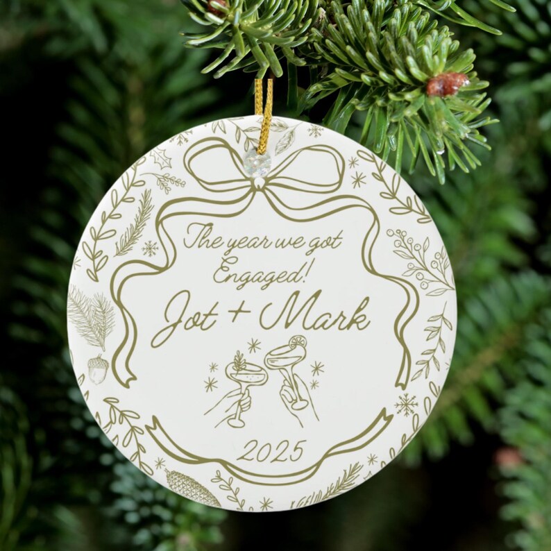 A round, white ceramic ornament with gold text and illustrations, reading "The year we got Engaged! Jot + Mark 2025," hangs on a green Christmas tree branch.