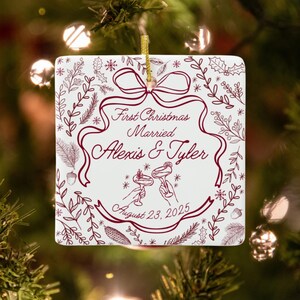 A square, white ceramic ornament with red text and illustrations, reading "First Christmas Married Alexis & Tyler August 23, 2025," hangs on a Christmas tree with blurred lights.