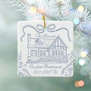 A square, white ceramic ornament with blue text and an illustration of a house, reading "The Burners Our first Christmas at 1827 N 60th St.," hangs on a light blue Christmas tree with blurred lights.