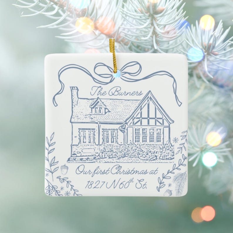 A square, white ceramic ornament with blue text and an illustration of a house, reading "The Burners Our first Christmas at 1827 N 60th St.," hangs on a light blue Christmas tree with blurred lights.