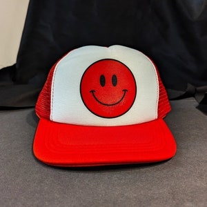 Könnte beinhalten: Eine rote und weiße Truckermütze mit einem roten Smiley auf der Vorderseite.