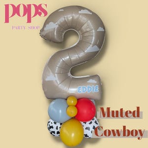 May include: A large number 2 balloon in a muted brown colour with white cloud accents, above a cluster of colourful balloons. The balloons include red, yellow, blue, and cow-print balloons. The name "Eddie" is printed on the number 2 balloon.
