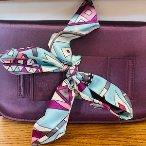 May include: A purple satin clutch with a colorful patterned scarf tied around the handle.