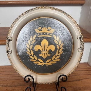 May include: Round decorative tray with a cream-colored rim and a dark gray center featuring a gold fleur-de-lis design. The tray has gold accents and is supported by a black metal stand. The tray has a diameter of approximately 12 inches.