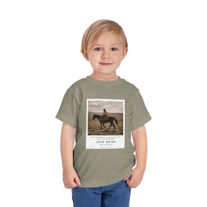 May include: A heather green toddler t-shirt with a framed image of a cowboy on horseback. The image includes text that reads "JOHN WAYNE THE COWBOYS". The shirt is made of a soft material.