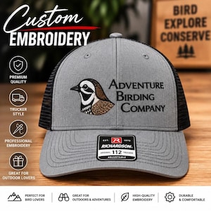 May include: A gray and black trucker hat featuring an embroidered quail graphic and the text Adventure Birding Company. The front panel is gray with a black mesh back, and a black Richardson 112 adjustable label is centered on the brim.