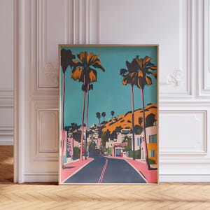 May include: A framed print of a colorful illustration of a street lined with palm trees and houses. The street is a vibrant pink and the houses are a mix of pink and yellow. The sky is a bright blue and the palm trees are a mix of green and yellow.