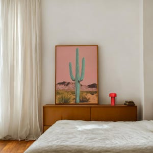 May include: A framed print of a large green cactus in a desert landscape with a pink sky. The print is hanging above a wooden dresser with a red lamp and books on top.