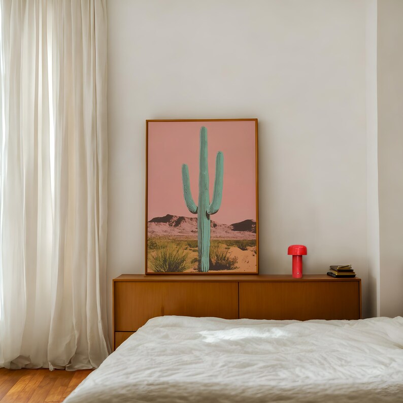 May include: A framed print of a large green cactus in a desert landscape with a pink sky. The print is hanging above a wooden dresser with a red lamp and books on top.